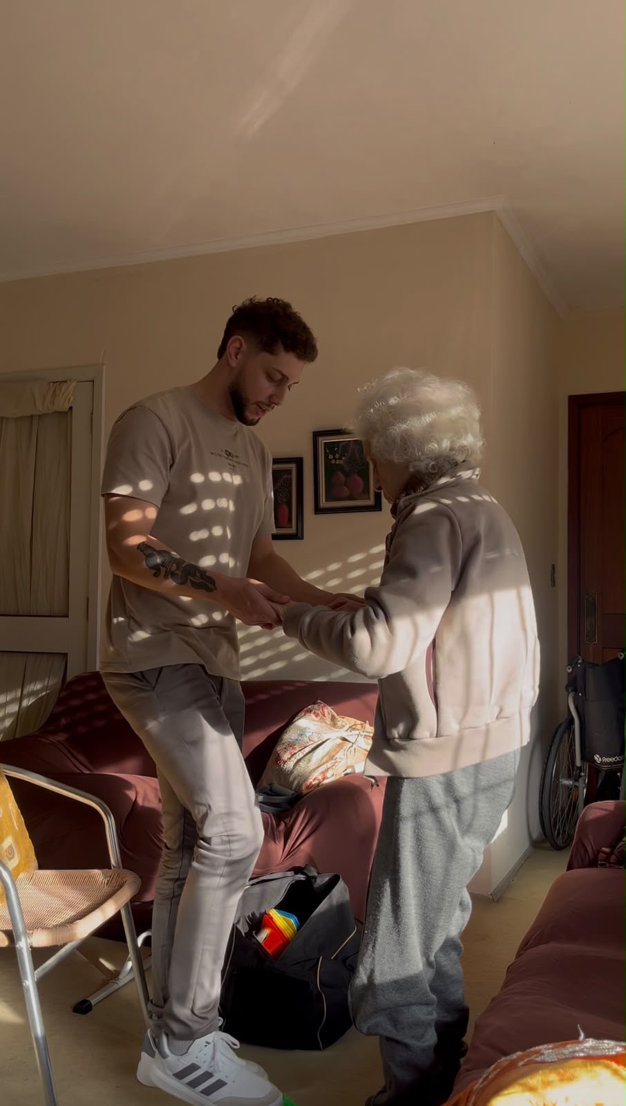 Arthur acompanha uma paciente idosa em pé, segurando suas mãos na sala de casa.