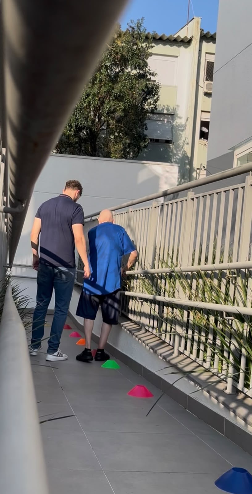 Arthur acompanha um paciente caminhando entre cones em uma passagem com corrimão.